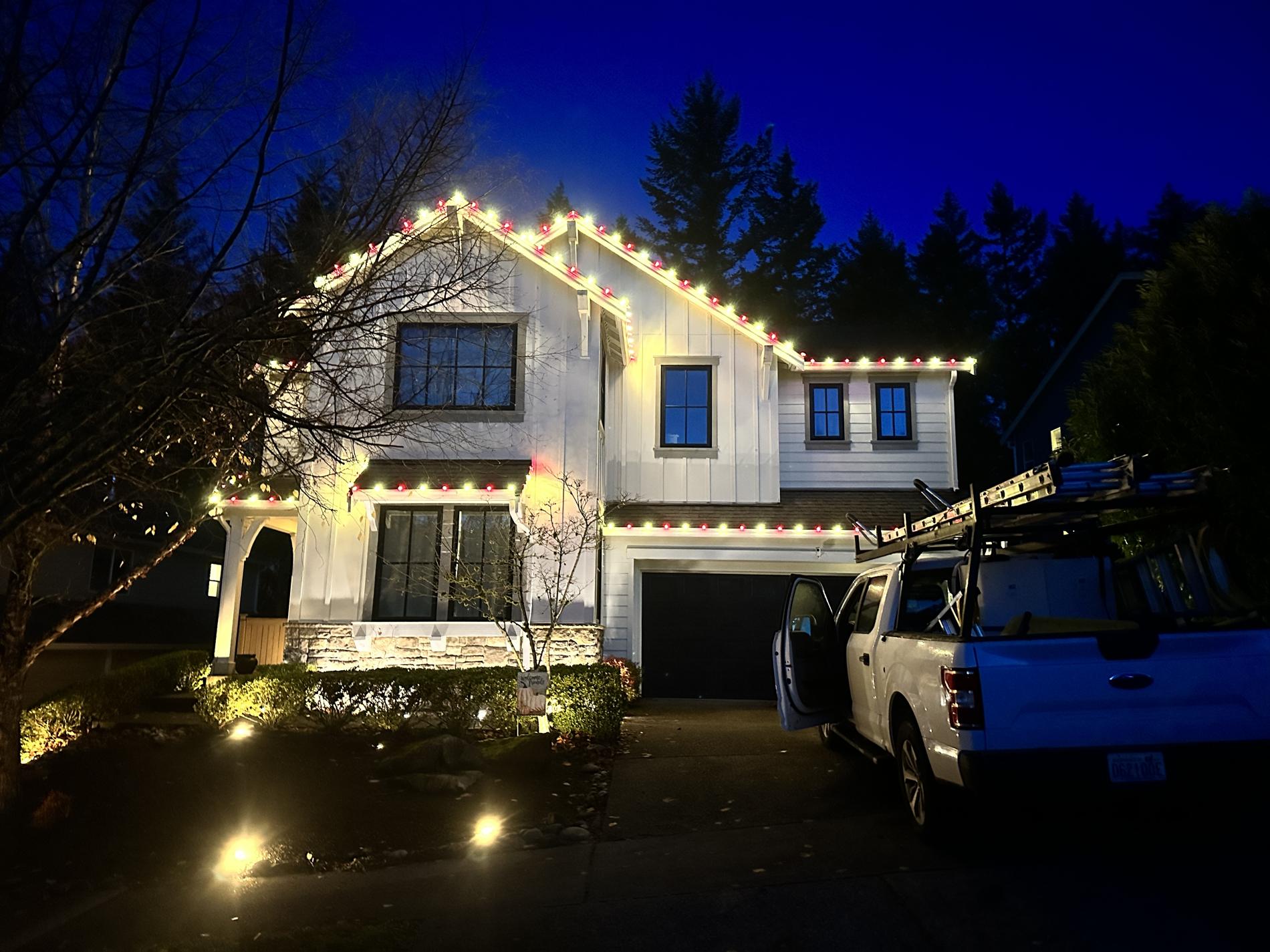 White farmhouse with red and warm white Christmas lights at dusk